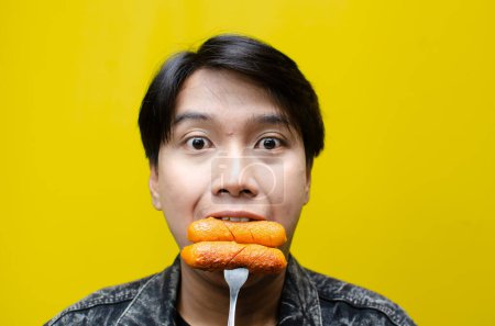 Téléchargez les photos : Extatique et expressif asiatique homme mange et mord saucisse à l'aide d'une fourchette isolée sur fond jaune - en image libre de droit