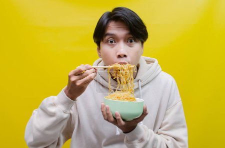Téléchargez les photos : Homme asiatique avec la bouche pleine de nouilles isolées sur fond jaune. un homme asiatique dans un sweat à capuche beige mange des nouilles instantanées avec faim en utilisant des baguettes et un bol. - en image libre de droit