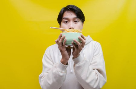 Téléchargez les photos : Gros plan, Asiatique en sweat à capuche beige mange des nouilles instantanées à l'aide de baguettes et d'un bol, isolé sur un fond jaune. - en image libre de droit
