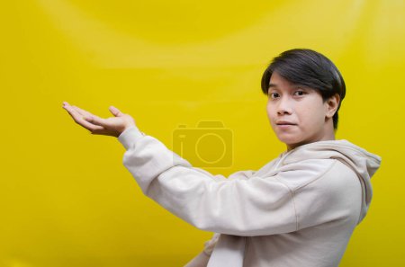 Photo for Side view of Asian man in a beige hoodie pointing his fingers and opening arms to up promote and advertise something good. - Royalty Free Image