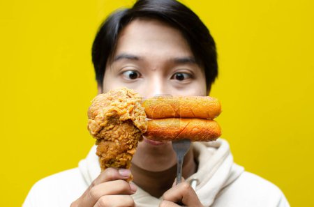 Photo for Happy surprised asian man holds fried chicken legs and sausages with wow expression isolated over yellow background. unhealthy fast food concept. food sale promotion - Royalty Free Image