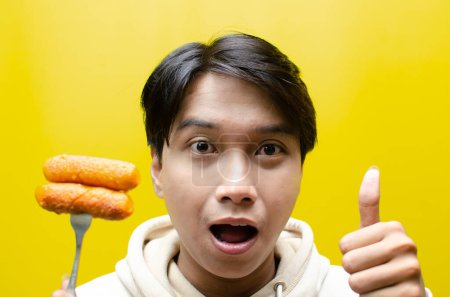 Foto de El hombre asiático come y sostiene salchichas sobre un tenedor aislado sobre fondo amarillo. concepto de comida rápida poco saludable. - Imagen libre de derechos