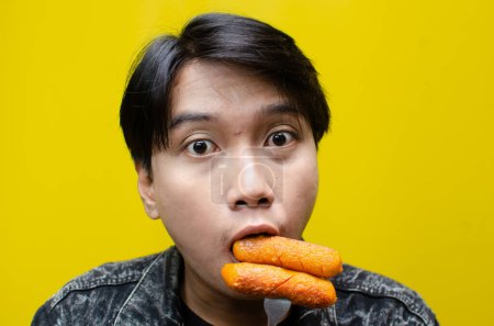 Foto de Extático y expresivo asiático hombre come y muerde salchicha usando tenedor aislado sobre amarillo fondo - Imagen libre de derechos