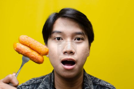 Téléchargez les photos : L'homme asiatique mange et tient des saucisses sur une fourchette isolée sur fond jaune. concept de restauration rapide malsaine. - en image libre de droit