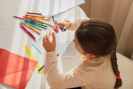Foto de Enthusiastic child in the room at the table cuts out his crafts from colorful colored paper with scissors in sunlight - Imagen libre de derechos
