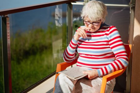 Foto de Senior woman in glasses sitting on a balcony near the sea and looking at magazine, solving a crossword - Imagen libre de derechos