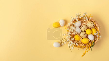 Téléchargez les photos : Easter candy chocolate eggs and almond sweets lying in a birds nest decorated with flowers and feathers on a yellow background. Top view long banner. Happy Easter concept. - en image libre de droit