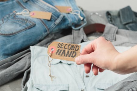 Photo for Clothes from females wardrobe on shelf of second hand store. Womans hand holding craft paper tag with inscription second hand. Circular fashion, eco friendly shopping, thrift stores concept - Royalty Free Image