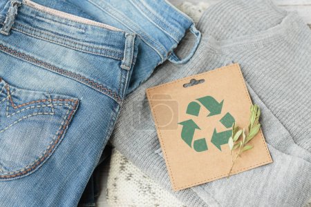 Nachhaltiges Stillleben mit Blue-Denim-Jeans, grauem Pullover und Bastelpapierkarte mit Recycling-Symbol. Second-Hand-Bekleidung Idee. Kreisförmige Mode, Spende, Charity-Konzept. Ansicht von oben