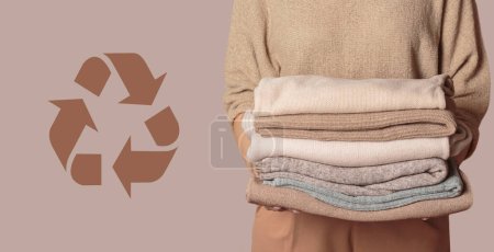 Photo for Woman holding stack of clothes with used wardrobe for reuse and circular economy logo on beige background. Reusing, recycling materials and reducing waste in fashion, second hand apparel idea - Royalty Free Image