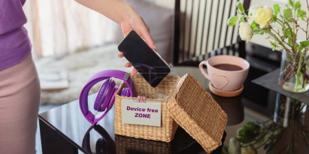 Photo for Close up of hand holding smartphone in wicker basket with inscription Device free zone. Woman putting phone into box with different gadgets at home. Digital detox and technology dependance concept - Royalty Free Image