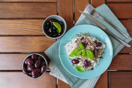 Feta-Käse auf blauem Teller mit Sesam und Leinsamen, serviert mit schwarzen Oliven und Basilikumblättern. Leckeres und gesundes Frühstück auf der Terrasse