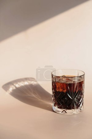 Whiskey cola cocktail with strong alcohol and ice in highball glass on light beige background with shadows and fantastic highlights and reflecting bright sunlight in daytime