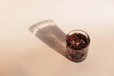 Whiskey cola cocktail with strong alcohol and ice in highball glass on light beige background with shadows and fantastic highlights and reflecting bright sunlight in daytime