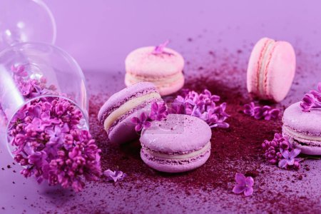 Pastel colored sweet french macaroons with lilac flowers and splash of dry blueberry powder on purple background. Beautiful composition for bakery and pastry shop