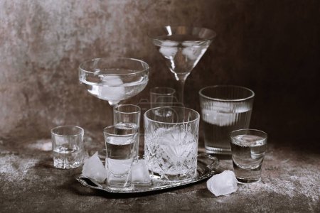 Collection of various glasses on silver tray filled with clear cold drinks with ice on grunge wall background