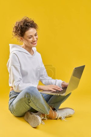 Téléchargez les photos : Vue latérale de la fille assise sur les jambes, travaillant à l'ordinateur portable. Jolie jeune femme aux cheveux bouclés souriant, regardant l'écran, tapant, faisant défiler. Isolé sur fond jaune studio. - en image libre de droit