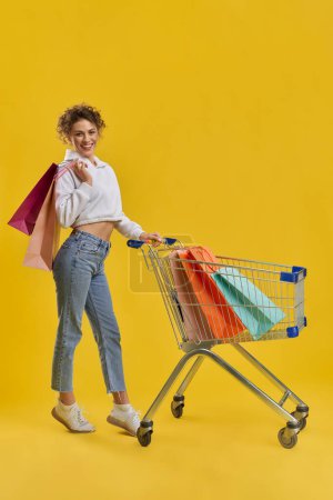 Side view of slim girl with blonde curly hair doing shopping. Pretty young woman standing on toes, driving cart, holding packages, looking at camera. Concept of urban life.