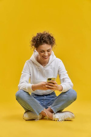 Photo for Front view of girl sitting in lotos pose, using smatphone. Beautiful female with curly hair smiling, looking at cell phone, chattig, scrolling. Concept of modern technologies. - Royalty Free Image