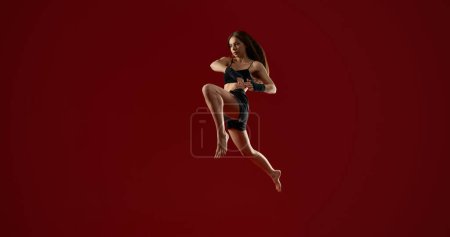 Widok z boku brunetka wysportowana kobieta z długimi włosami skacząc wysoko, na czerwonym tle studio. Dość silny trening kobiet w powietrzu. Pojęcie sztuk walki.