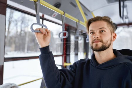 Téléchargez les photos : Front view of young male with beard going to work by public transport, standing on bus, waiting. Brunette man holding handle, wearing black khudi. Concept of everyday routine. - en image libre de droit