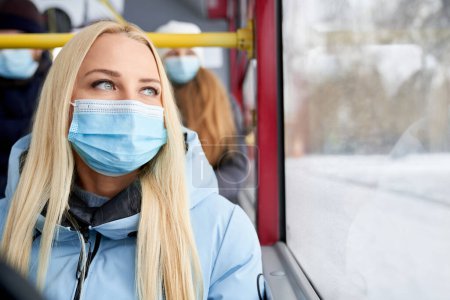Photo for Front view of blonde, attractive woman going home by public transport, wearing protecting mask. Passengers sitting on bus, protecting from coronavirus. Concept of everyday routine. - Royalty Free Image