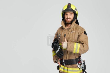 Photo for Happy caucasian firefighter in uniform showing thumb up in studio. Front view of confident bearded fireman giving like, while smiling to camera, on gray background. Concept of work, hand gesture. - Royalty Free Image