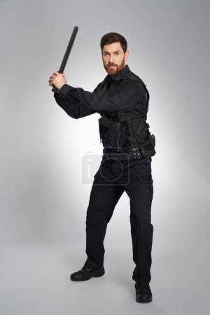 Photo for Focused caucasian police officer brandishing with police baton. Front view of dark-haired guy in police uniform beating, attacking camera, on gray background. Concept of police work, equipment. - Royalty Free Image