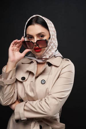 Téléchargez les photos : Élégante dame caucasienne regardant au-dessus des lunettes de soleil à la caméra à l'intérieur. Portrait de femme sérieuse à la mode portant un trench-coat et un foulard droit, isolé sur noir. Mode, concept de style. - en image libre de droit