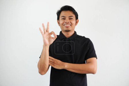 Téléchargez les photos : Jeune asiatique homme souriant amical tout en donnant OK doigt signe portant noir polo t-shirt isolé sur fond blanc - en image libre de droit