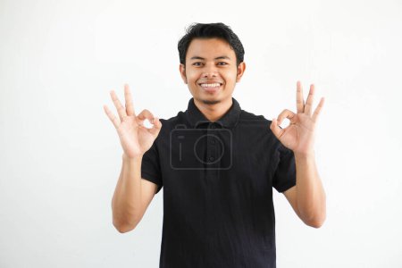Téléchargez les photos : Jeune asiatique homme souriant amical tout en donnant OK doigt signe portant noir polo t-shirt isolé sur fond blanc - en image libre de droit