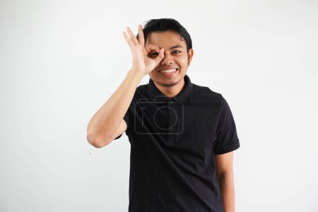 Téléchargez les photos : Jeune asiatique homme excité garder ok geste sur oeil, portant noir polo t-shirt isolé sur fond blanc - en image libre de droit