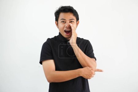 Photo for Young asian man posing on a white backdrop saying a gossip, pointing to side reporting something, wearing black polo t shirt. - Royalty Free Image