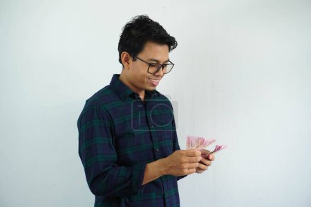 Téléchargez les photos : Jeune asiatique homme souriant heureux en comptant l'argent qu'il détient isolé sur fond blanc - en image libre de droit