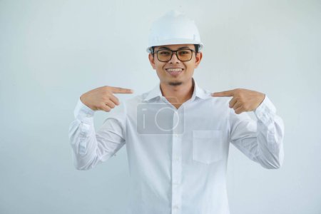 Téléchargez les photos : Jeune architecte asiatique homme portant le casque de sécurité constructeur souriant avec le visage heureux regardant et le pointant lui-même isolé sur fond blanc - en image libre de droit