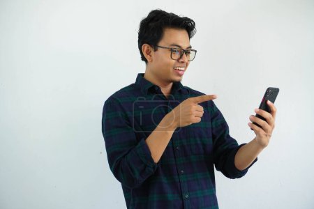 Foto de Sonriendo joven asiático hombre cuando mirando a su teléfono móvil y punto con un dedo aislado sobre fondo blanco - Imagen libre de derechos
