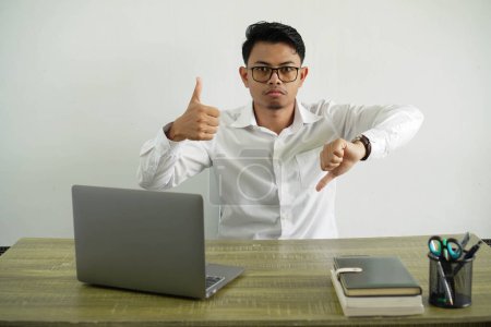 Téléchargez les photos : Jeune homme d'affaires asiatique dans un lieu de travail faisant signe bon-mauvais. indécis entre oui et non, portant une chemise blanche avec des lunettes isolées - en image libre de droit