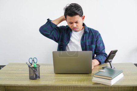 An Asian male freelance worker is sitting looking at a laptop showing a confused gesture with his hands holding the back of his head