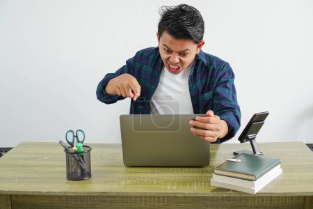 wütender asiatischer Geschäftsmann sitzt an seinem Arbeitsplatz und zeigt gebrüllt auf den Laptop-Bildschirm