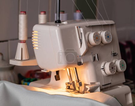 Foto de Una máquina de coser blanca con cuatro hilos de carrete y una lámpara en el escritorio. Overlock blanco. Concepto de lugar de trabajo. - Imagen libre de derechos