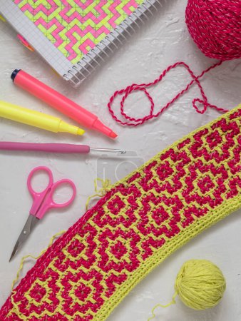Foto de Yellow pink crochet fabric with mosaic pattern. Workplace with crochet fabric, yarn balls, crochet hook, notebook and markers. Handmade concept. - Imagen libre de derechos