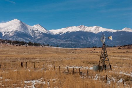Téléchargez les photos : Un vieux moulin à vent se dresse au-dessus des terres du ranch dans la vallée humide de montagne sous les montagnes de Sangre de Cristo couvertes de neige du Colorado. - en image libre de droit