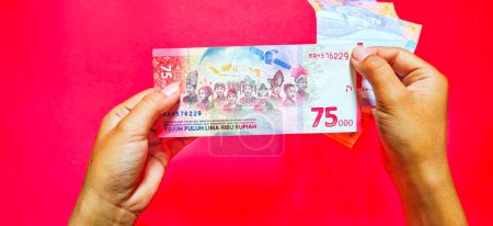 Téléchargez les photos : Homme détenant un nouveau billet émis en 2022 Rp.75.000. Monnaie rupiah isolée sur fond rouge - en image libre de droit