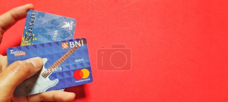 Téléchargez les photos : Jakarta, Indonésie - 29 janvier 2023. Homme détenant deux cartes bancaires indonésiennes, à savoir BRI et BNI. Concept d'entreprise isolé sur fond rouge. - en image libre de droit