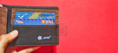 Photo for Jakarta, Indonesia - January 29, 2023. Man holding a wallet containing two Indonesian bank cards, namely BNI and BRI. Business concept isolated on red background. - Royalty Free Image