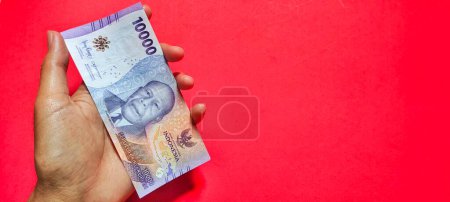 Téléchargez les photos : Homme tenant de nouveaux billets de banque roupies rp10000 isolés sur un fond rouge - en image libre de droit