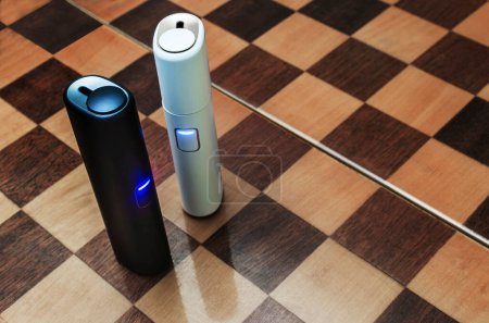 Tecnología de cigarrillos electrónicos. Vista superior de dos cigarrillos híbridos eléctricos sistema de calefacción de tabaco en carcasa blanca y azul en un tablero de ajedrez