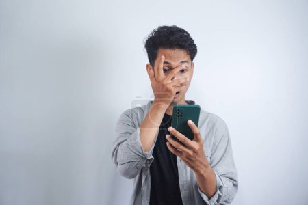 Photo for Close-up portrait of shocked, scared asian man closing one eye while looking at cell phone isolated on white background - Royalty Free Image