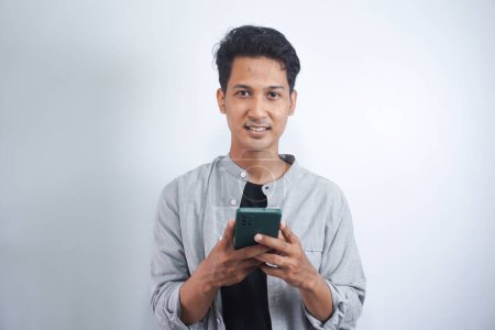 Photo for Positive Asian guy, wearing a basic shirt, using his smartphone, typing a message, browsing the internet, social media, stands on isolated white background, looking at camera, smiling - Royalty Free Image