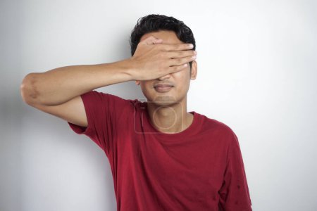 Handsome Asian man closing his eyes with one hand wearing red t-shirt isolated on white background
